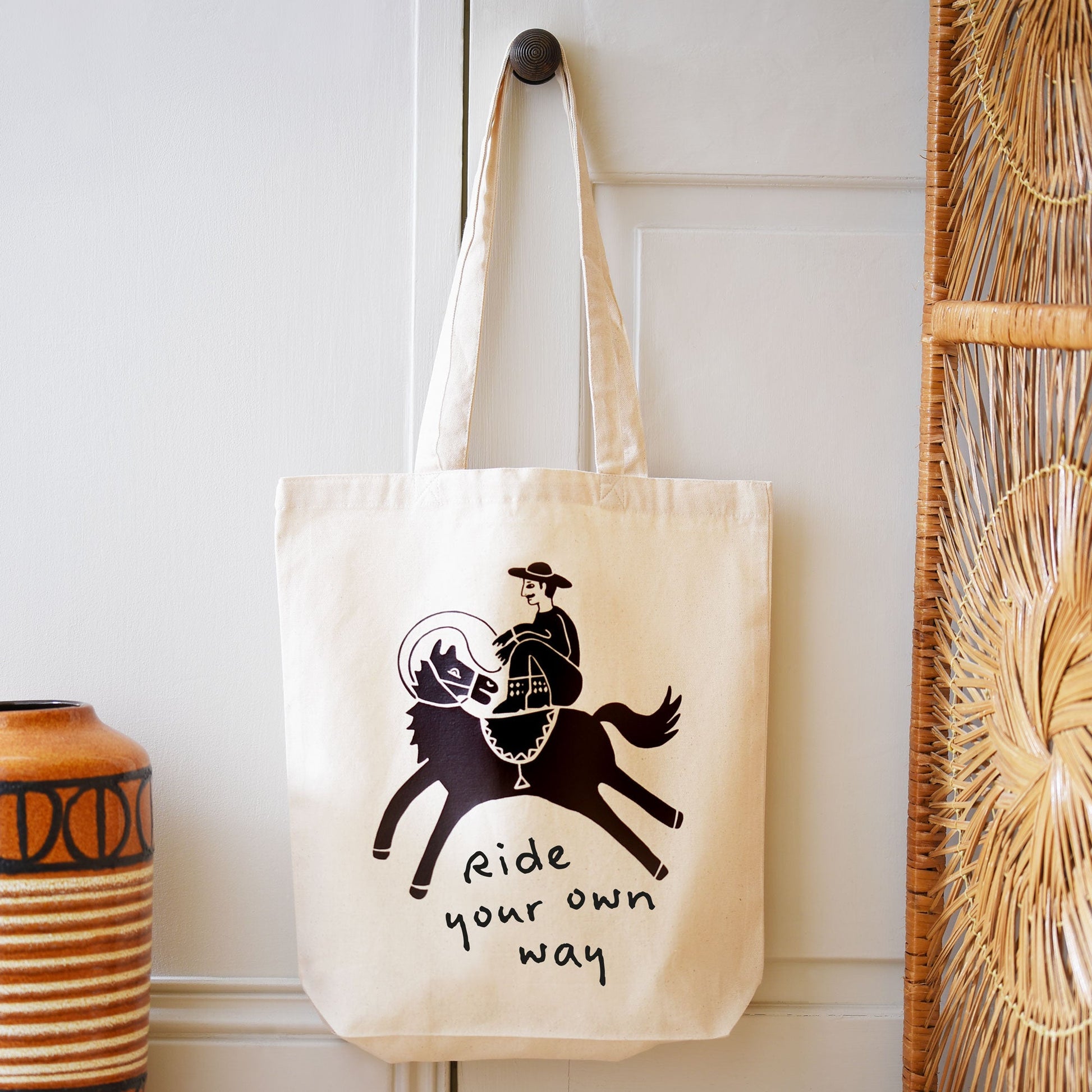 A cotton canvas shopping tote bag with an illustration of a cowboy riding a horse and the phrase 'ride your own way' printed on the front.