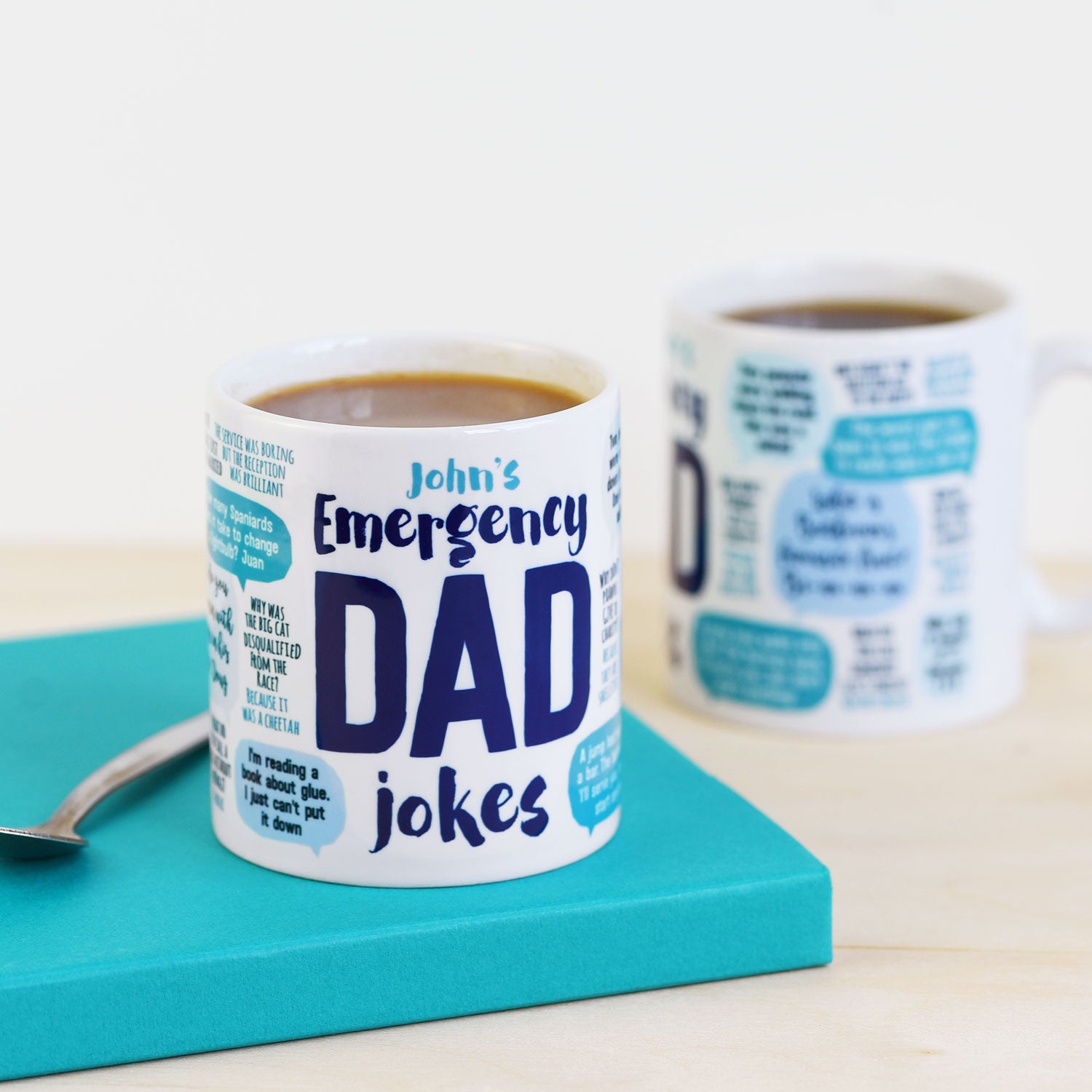 Mug with 'John's Emergency Dad Jokes' text on a teal coaster with coffee.