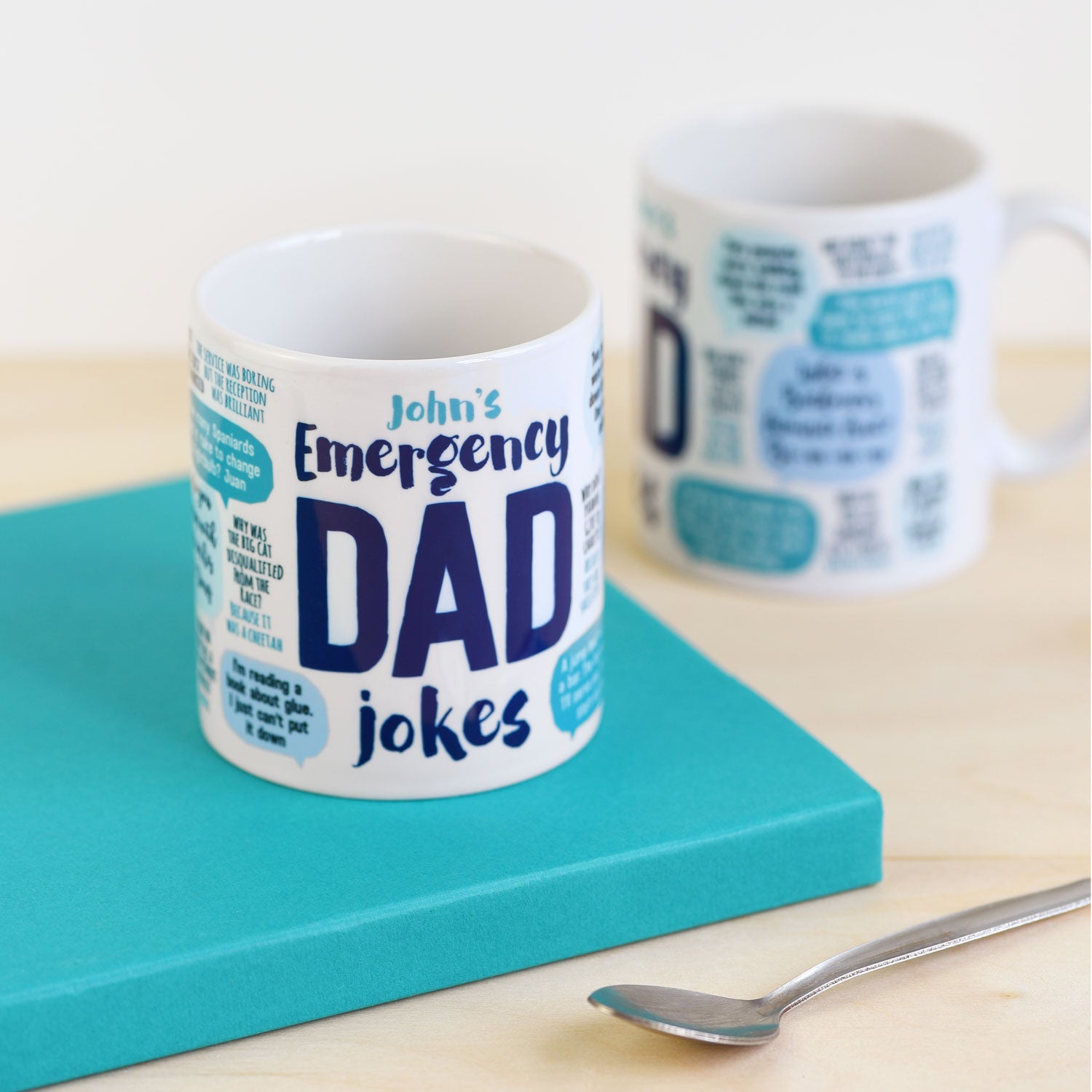 Mug with 'John's Emergency Dad Jokes' text on a turquoise book with a spoon beside it.